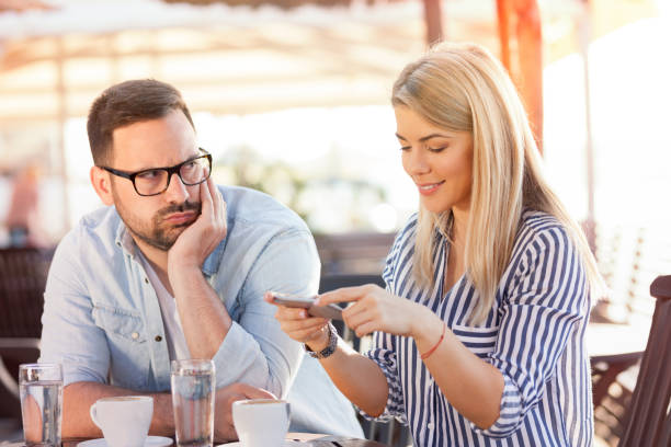 Young woman using phone while sitting at cafe with her boyfriend Internet addiction. Attractive woman messaging online via social networks while sitting at cafe with her boyfriend who is having unhappy and bored look couple on bad date stock pictures, royalty-free photos & images