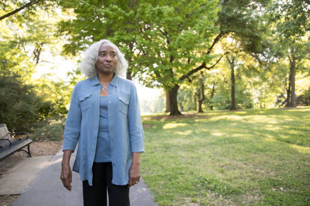 A senior woman with gray and white hair looks away from the camera.  She has a serious expression and is standing in the park.  She is wearing a blue shirt and undershirt and black slacks.  She is African ethnicity.  Outdoors, lens flare.