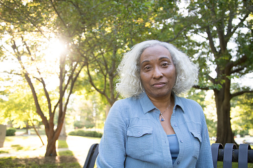 A senior woman with gray and white hair on a bench in the park.  She's looking at the camera with a serious expression.  She is wearing a blue shirt and undershirt.  She is African ethnicity.