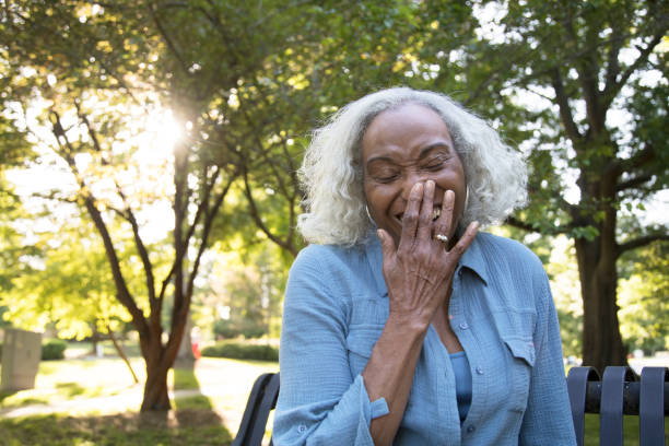 Senior Woman Outside on a Bench Laughing A senior woman with gray and white hair on a bench in the park.  She's laughing and holding her hand up to cover her face.  She is wearing a blue shirt and undershirt.  She is African ethnicity. senior adult women park bench 70s stock pictures, royalty-free photos & images