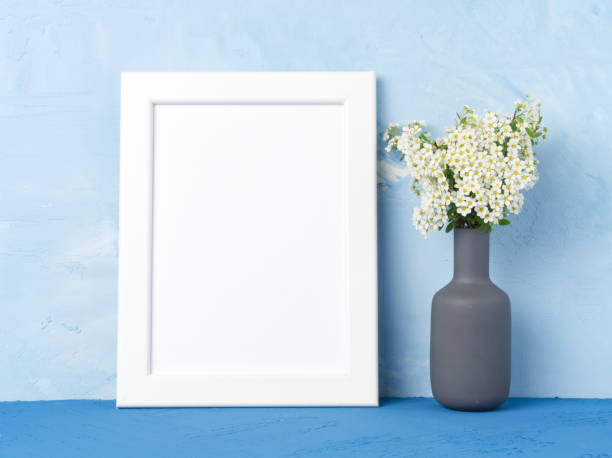 istock Blank white frame, flower in vase on dark blue table against blue concrete wall with copy space. Mock up. 962962366