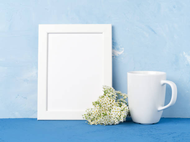 istock White mug with tea or coffee, frame, flower bouquet on blue table opposite blue concrete wall. Mock up with empty space 962964768
