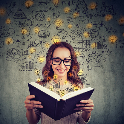 Young girl in glasses reading big book and having plenty of successful ideas looking happy on gray background.