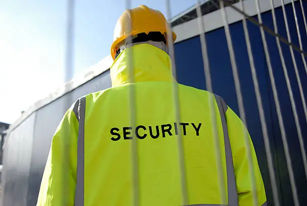 Low view of a security man behind a wire fence wearing a hardhat on a construction site.