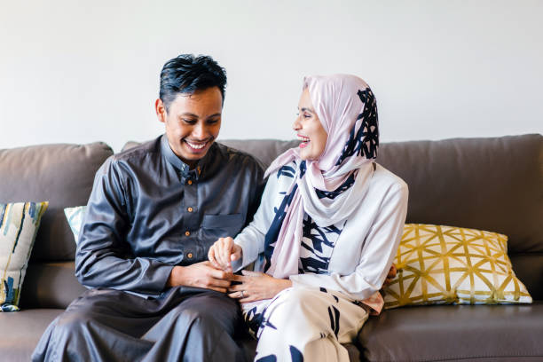 Portrait of a young married Muslim couple sitting on the couch. They are tickling one another in joy during Ramadan. They are both laughing candidly as they sit and tickle one another. They are dressed in traditional garb (the woman is in a hijab headscarf) to celebrate Ramadan. happy malay couple stock pictures, royalty-free photos & images