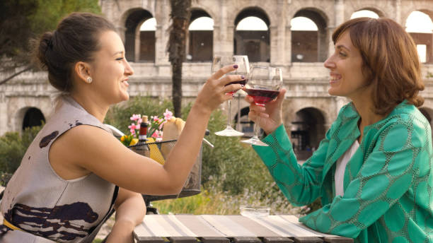 dos turistas de feliz joven mujer sentada en la mesa de un bar restaurante frente del coliseo en roma bebida y brindis con una copa de vino tinto italiana. vestido con estilo colorido en un día de verano al atardecer - rome cafe art italy fotografías e imágenes de stock