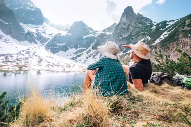 Father and son backpackers sit near the mountain lake and enjoy mountain snowy peaks