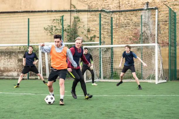 Young man wearing football bib with the ball as opponent comes in to tackle and goalkeeper watches