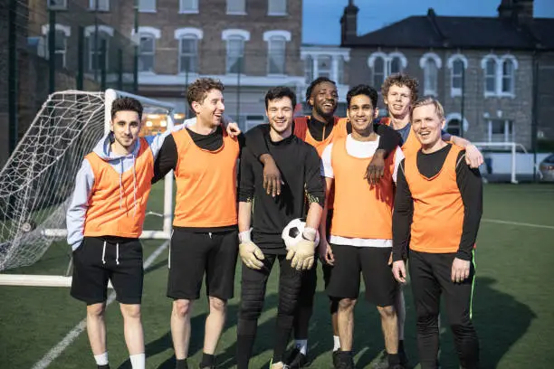 Group of friends on 7 a side football team standing by the goal and looking towards camera, goalkeeper in middle holding football