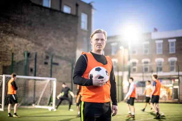 Mid adult man in orange bib on urban football pitch with team mates playing in the background