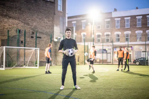 Floodlit urban football pitch at night, man in his 20s standing with ball and facing camera