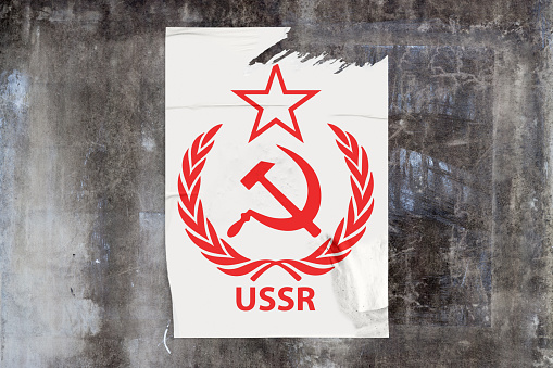 Full-frame weathered concrete wall with a torn poster in the middle depicting in red, the Soviet Communist USSR Laurel Hammer Sickle symbol with 