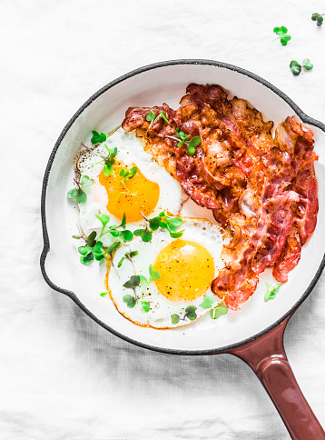 Classic Breakfast - fried eggs and bacon in a cast iron pan on a light background, top view