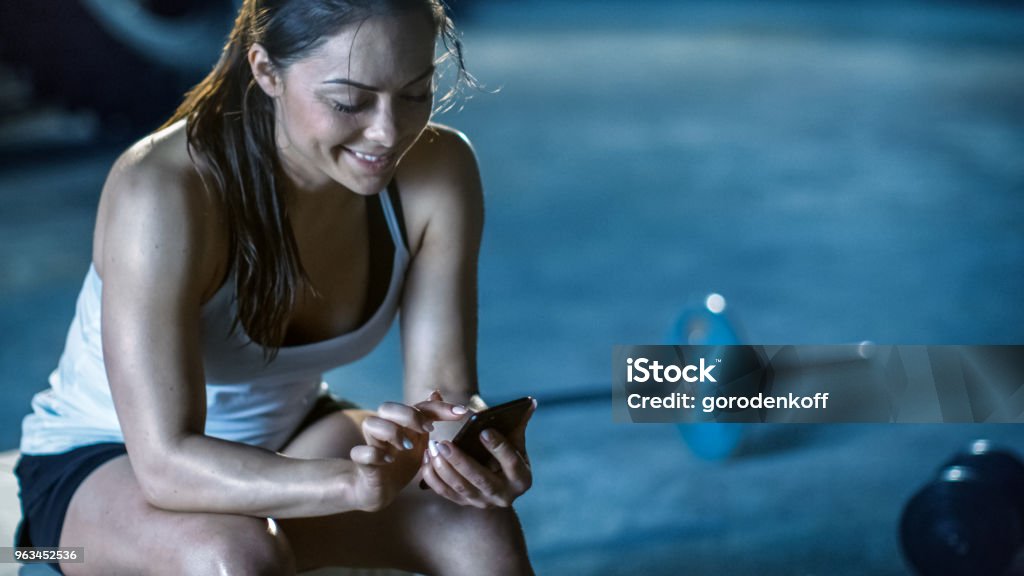 Schöne sportliche Brünette nutzt Smartphone während der Ruhepause auf der Bank nach ihrer Intensivtraining der Bodybuilding-Fitness-Studio. - Lizenzfrei Trainingsraum - Freizeiteinrichtung Stock-Foto