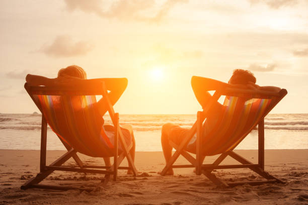 Romantic holiday travel. Silhouette of happy young couple sitting in deck chairs in luxury beach hotel at sunset near the sea. Love and relationship concept. Summer vacation in tropical paradise island. stock photo