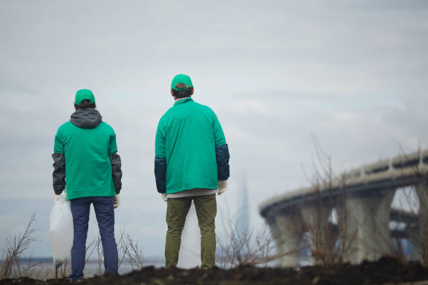 Two greenpeacers Backs of two guys in green uniform standing in front of littered territory against grey sky greenpeace activists stock pictures, royalty-free photos & images