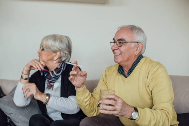 Elderly happy man smoking cigarette and holding drinking glass while his wife turning back at him