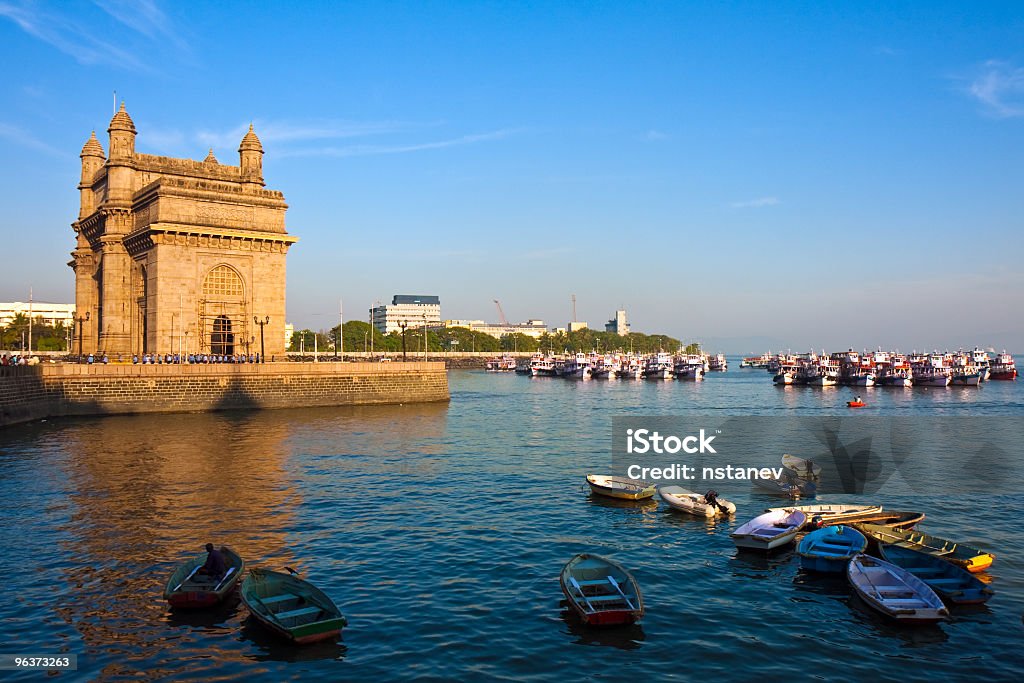 Gateway of India monument in Mumbai at sunset Gateway of India in Mumbai during sunset.  There are several small boats in blue water and a castle-like building towards the left.  There is land visible in the distance. Mumbai Stock Photo