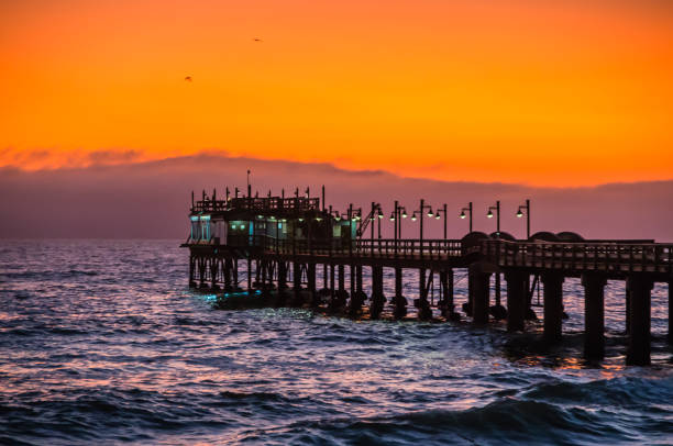 Wooden pier reaching out in the Atlantic ocean, with restaurant at the end, in front of orange and purple sky at sunset. Swakopmund, Namibia, Southern Africa.