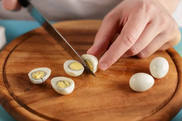 Man cuts boiled quail eggs on wooden cutting board close-up. Step by step recipe of homemade tuna salad.