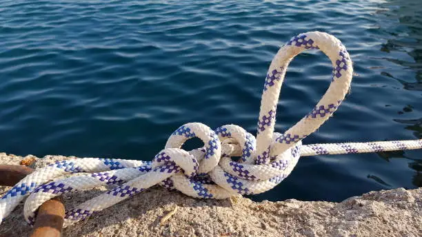 Marine knot on boat pier with deep blue sea waving water. White and blue nautical rope knot on the clear water surface background. Summer vacation and sea travel.