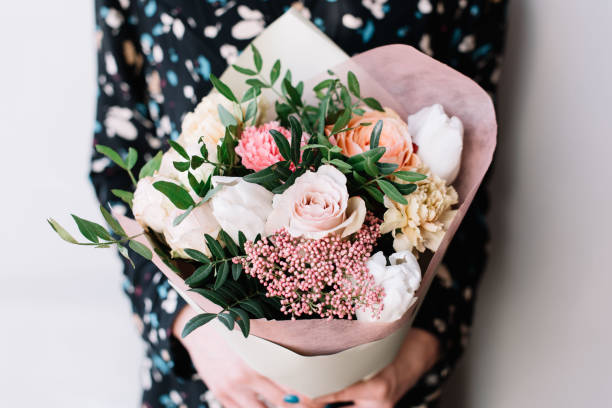 Very nice florist woman holding a beautiful colourful blossoming flower bouquet of fresh Quicksand roses, carnations, ranunculus, peony, pistachio leaves on the grey wall background stock photo