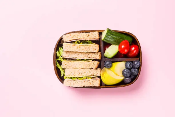 School wooden lunch box with sandwiches, vegetables, , tomatoes and fruits on pink background. Healthy children eating concept flat lay. Top view with copy space.