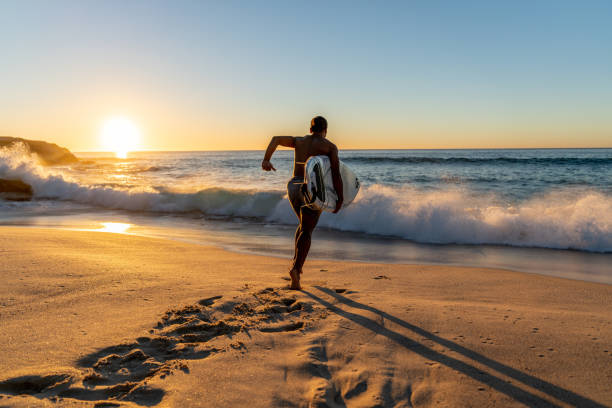 surfeur en cours d’exécution dans l’eau transportant son conseil d’administration - surfboard photos et images de collection