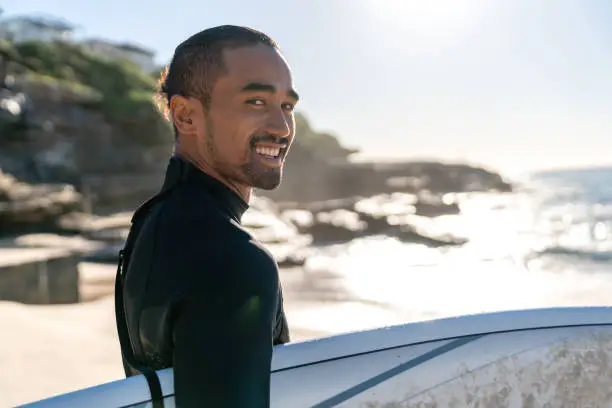 Portrait of a handsome surfer carrying his board and looking at the camera smiling - sports concepts