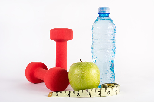 Red dumbbell, measuring tape, green apple and water bottle on white background in studio. Diet, fitness and sport concept