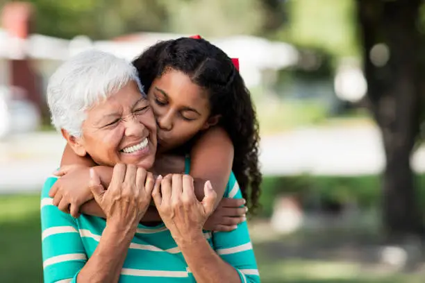 A loving teen girl embracing and kissing her happy grandmother from behind and holding each other outdoors.