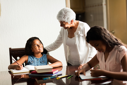 A latin senior woman standing next to her granddaughters at the table while they do their homework at home.