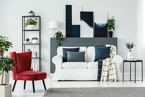 Red armchair, white, comfy sofa with blue pillows and plants in an elegant living room interior