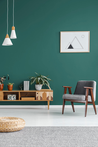 Simple geometric poster hanging on the wall in green living room interior with grey armchair, wooden cupboard and fresh plants