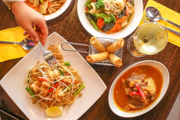 The image shows a hand with a fork reaching for the plate of Pad Thai prawns, a metal basket with 4 spring rolls, a red curry, a bowl of stir fried vegetables, and a glass of white wine.