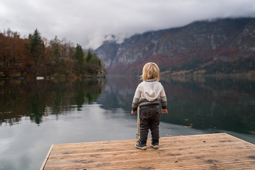 Back view of small kid standing on a pier by the lake Bohinj and looking at the view during an autumn day.