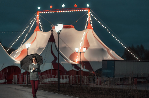 Beautiful young woman with long brown hair dressed in a casual clothing, she holds the black umbrella. The woman is walking the walkway near a white circus tent with a night illumination. She pensive looks down. Shooting in a spring evening