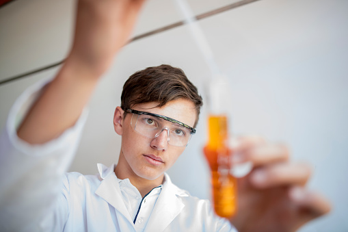 Male high school student dripping liquid into test tube. Teenage boy is doing science experiment in laboratory. He is wearing protective eyewear.