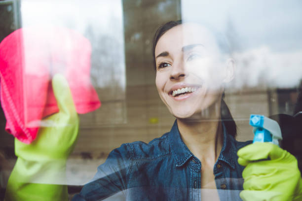 istock Beautiful Young Woman is Using a Duster and a Spray and Smiling While Cleaning Windows in the House 964208664
