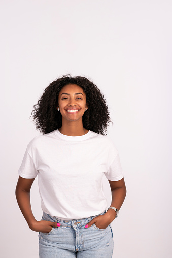 Smiling young woman with a natural look free from makeup looking at camera with a friendly smile. The woman wearing casual simple clothes: a white t-shirt and jeans. Waist up portrait of elated beautiful African female model on white background.