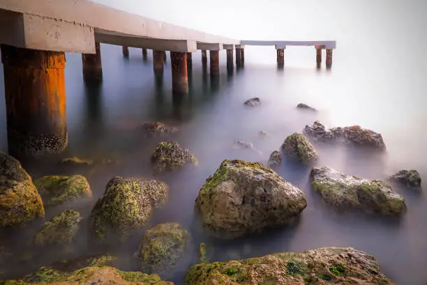 The pier is near the sea. It was shot as long exposure so there are a lot of rocks. Rocks has mosses. The sea looks as foggy.