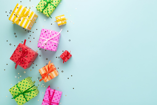 Holiday flat lay with gift boxes wrapped in colorful paper and tied with bows on blue background, decorated with confetti. Birthday, Christmas and sale concept, top view.