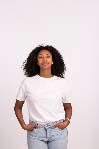 Beautiful young woman standing with hands in the pocket, looking at camera with a serious facial expression. The woman with a natural look, no makeup at all, wearing white t-shirt and jeans. Waist up portrait of elated beautiful African female model on white background.
