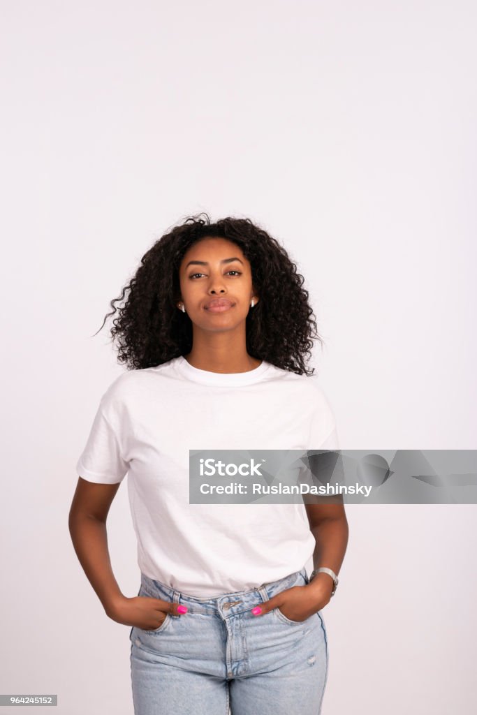 Woman standing with hands in the pockets. Beautiful young woman standing with hands in the pocket, looking at camera with a serious facial expression. The woman with a natural look, no makeup at all, wearing white t-shirt and jeans. Waist up portrait of elated beautiful African female model on white background. Women Stock Photo