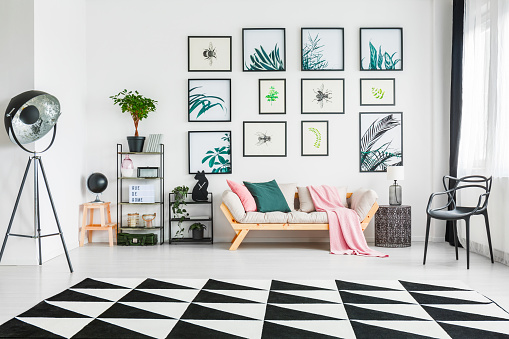Real photo of a couch with cushions and a blanket standing between the shelves with ornaments and a chair in a living room interior with painting on the wall, black lamp and black and white triangle rug