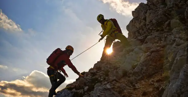Male mountaineer pulling his friend with a rope on mountain at sunset.