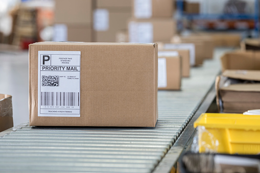 A brown cardboard box sits on a conveyor belt in a distribution warehouse. It has a priority mail label on it as well as a bar code and QR code.