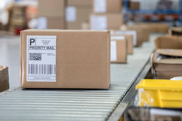 Cardboard box waiting to be shipped in distribution warehouse A brown cardboard box sits on a conveyor belt in a distribution warehouse. It has a priority mail label on it as well as a bar code and QR code. brown box stock pictures, royalty-free photos & images