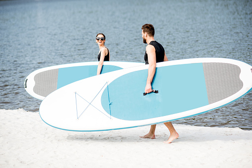 Beautiful couple in black swimwear walking with standup paddleboard on the beach ready for surfing