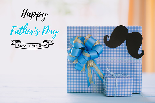 Happy fathers day concept. Stack of beautiful gift box with black mustache paper on bright blue pastel wooden table background.